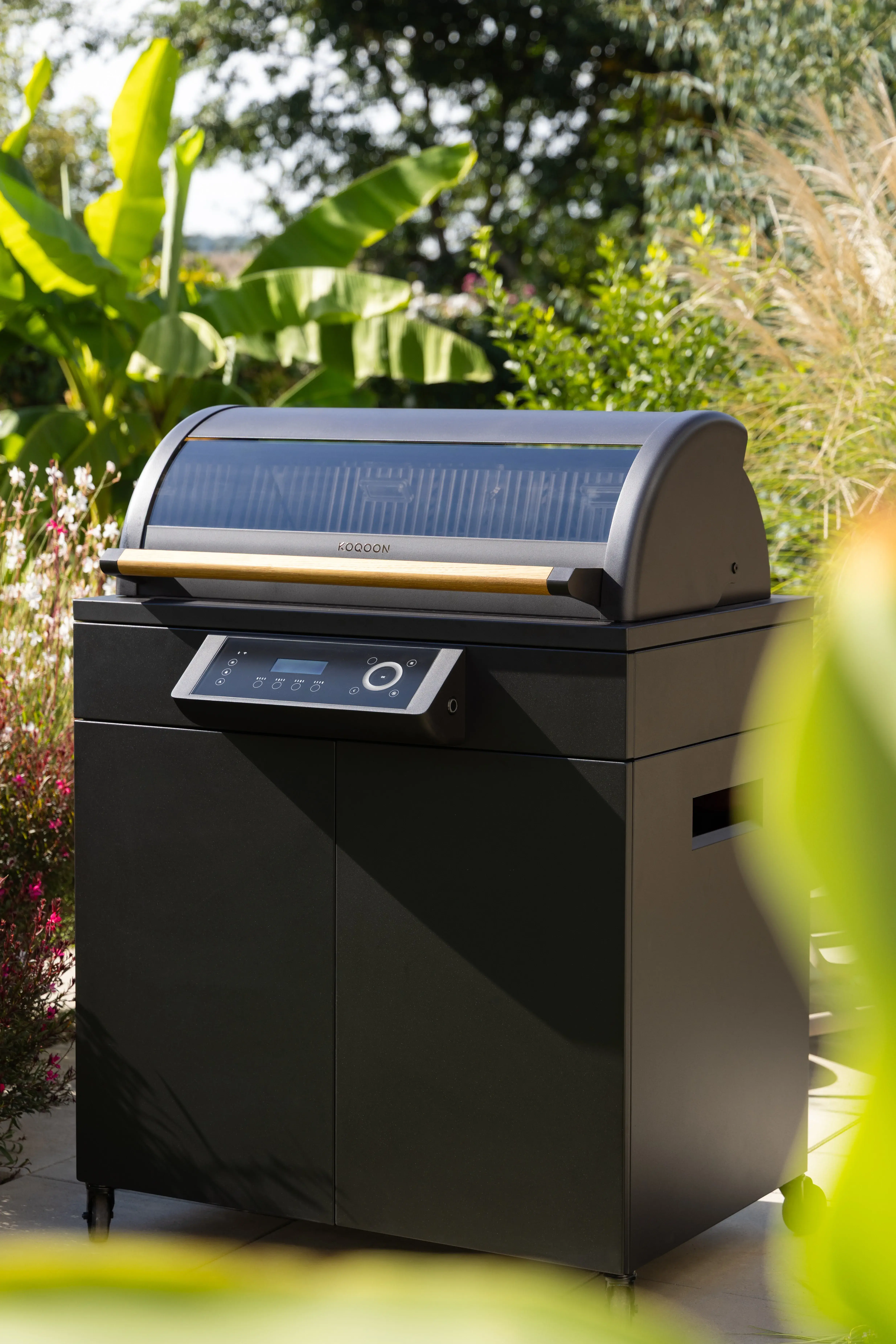 Schwarzer Grill im Garten, umgeben von grünen Pflanzen bei Sonnenschein.
