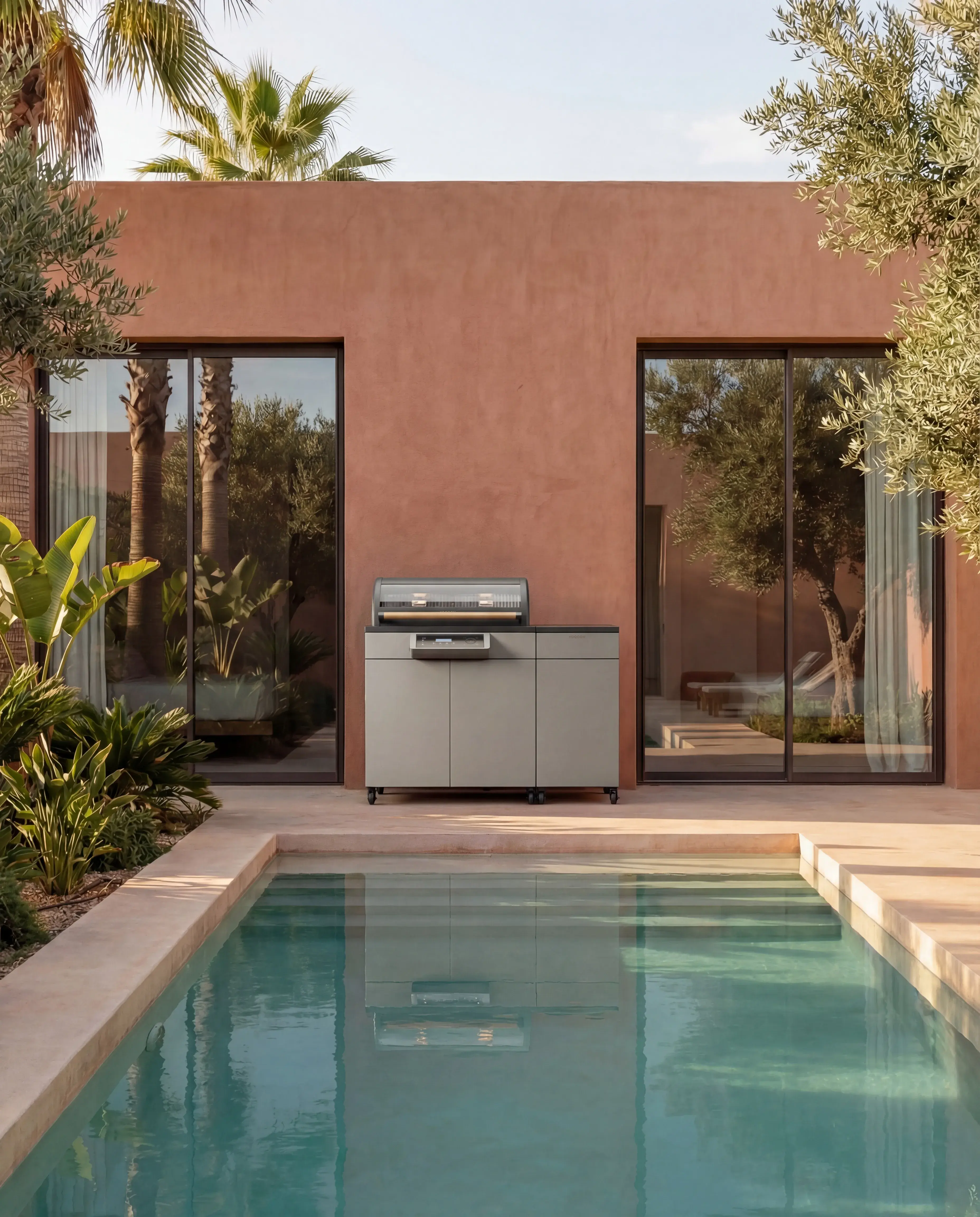 Moderner Grill auf Terrasse vor Pool und rosafarbenem Haus, umgeben von Palmen und Grün.
